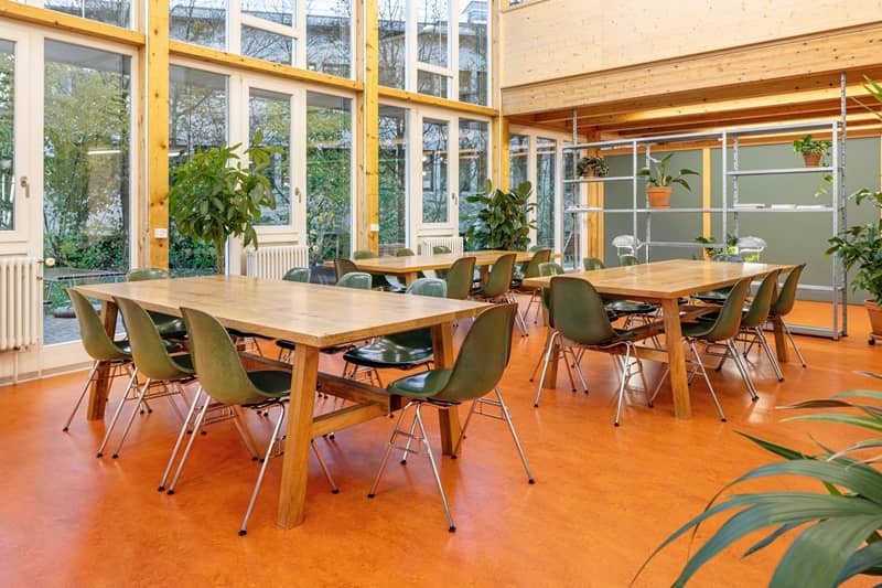 Eine Kantine mit h&ouml;lzernen Tischen, gr&uuml;nen St&uuml;hlen und viel Pflanzen